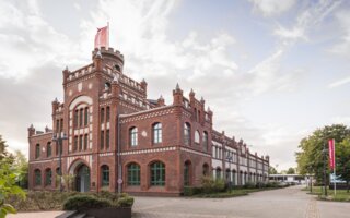 Das Bild zeigt ein historisches, rotes Backsteingebäude im neugotischen Stil, umgeben von Pflanzen und einem klaren Himmel mit Wolken.