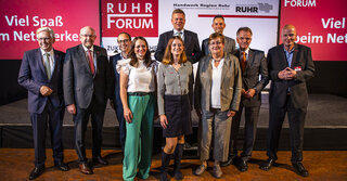 Das Bild zeigt eine Gruppe von Personen auf der Bühne eines Events. Sie lächeln und stehen vor einem Hintergrund mit dem Schriftzug "RUHR FORUM".