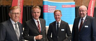 Das Bild zeigt vier Männer in Anzügen, die in einem modernen Raum stehen. Sie wirken freundlich und sind bereit für ein Gespräch oder eine Veranstaltung.