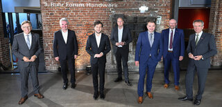 Eine Gruppe von sieben Männern in formeller Kleidung steht vor einer Backsteinwand. Sie wirken professionell und aufmerksam, bereit für eine Veranstaltung.