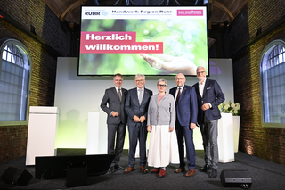 Das Bild zeigt fünf Personen auf einer Bühne mit einem Willkommen-Schild. Sie stehen lächelnd vor einem modernen, festlichen Hintergrund.